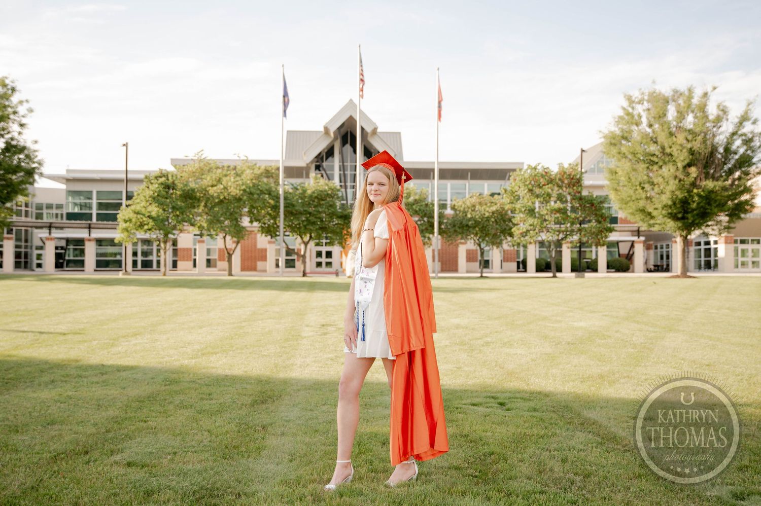 A graduate in white dress and bright orange cap and gown poses on a green lawn in front of a school building.