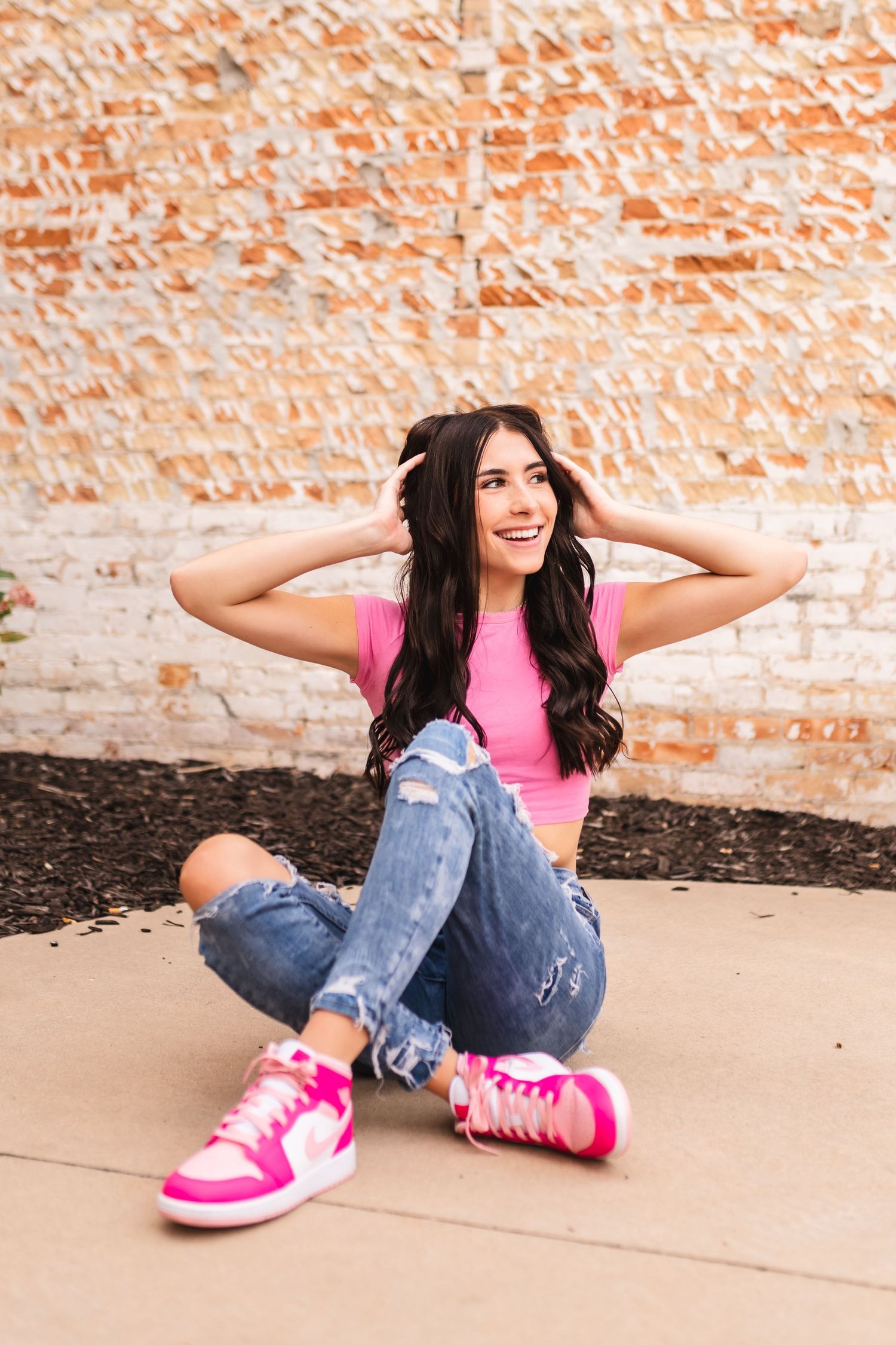 A senior girl sitting on the ground with her trademark pink shoes, playing with her hair.