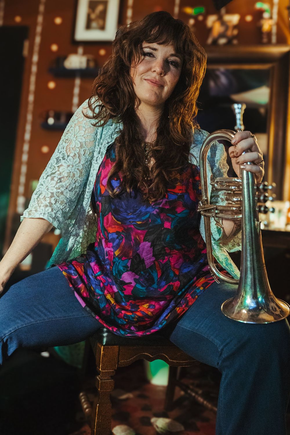 A musician in a floral dress and lace cardigan poses with a trumpet in a dimly lit venue.