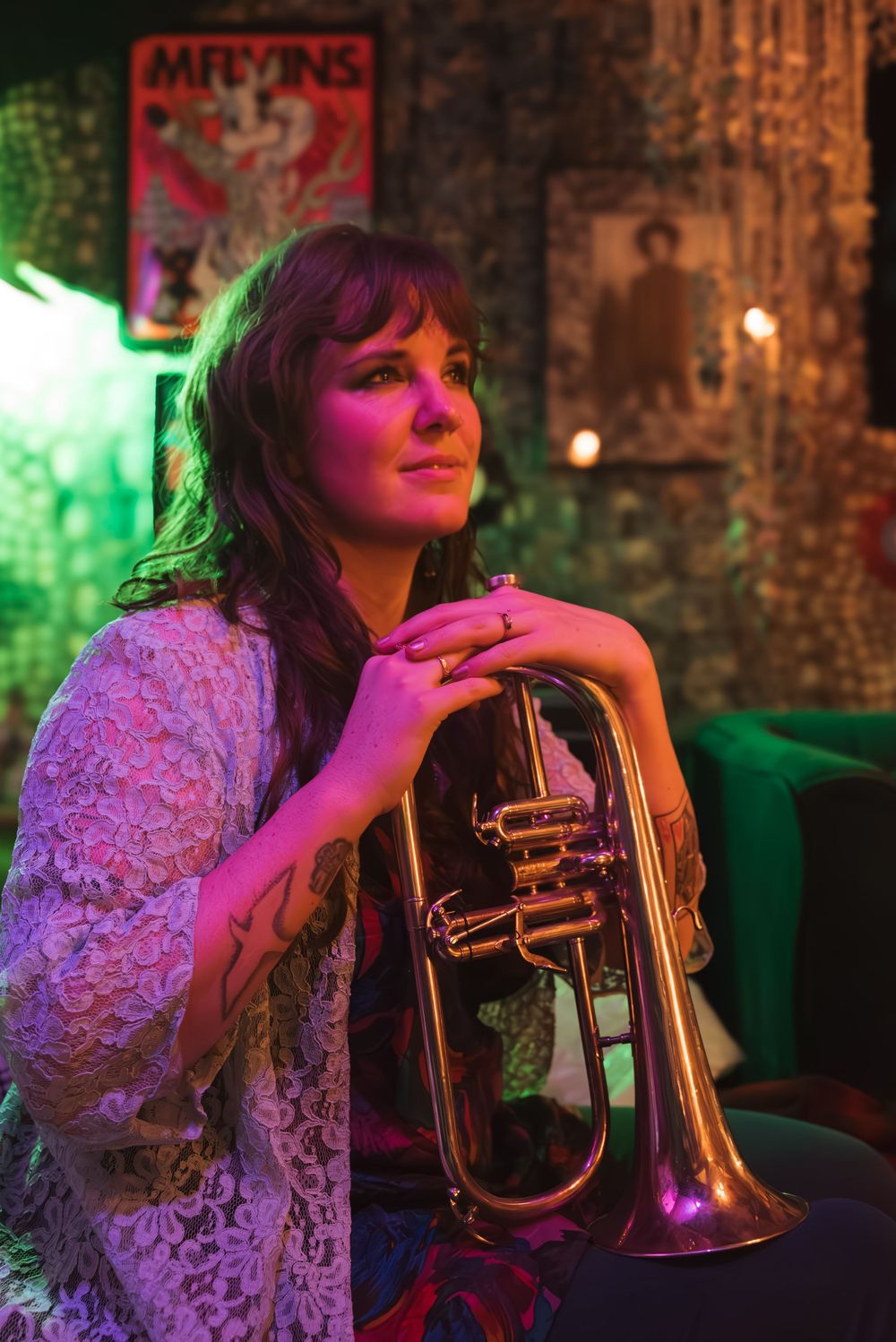 Musician in white lace top illuminated by vibrant purple and green lights holds flugelhorn in moody venue setting.