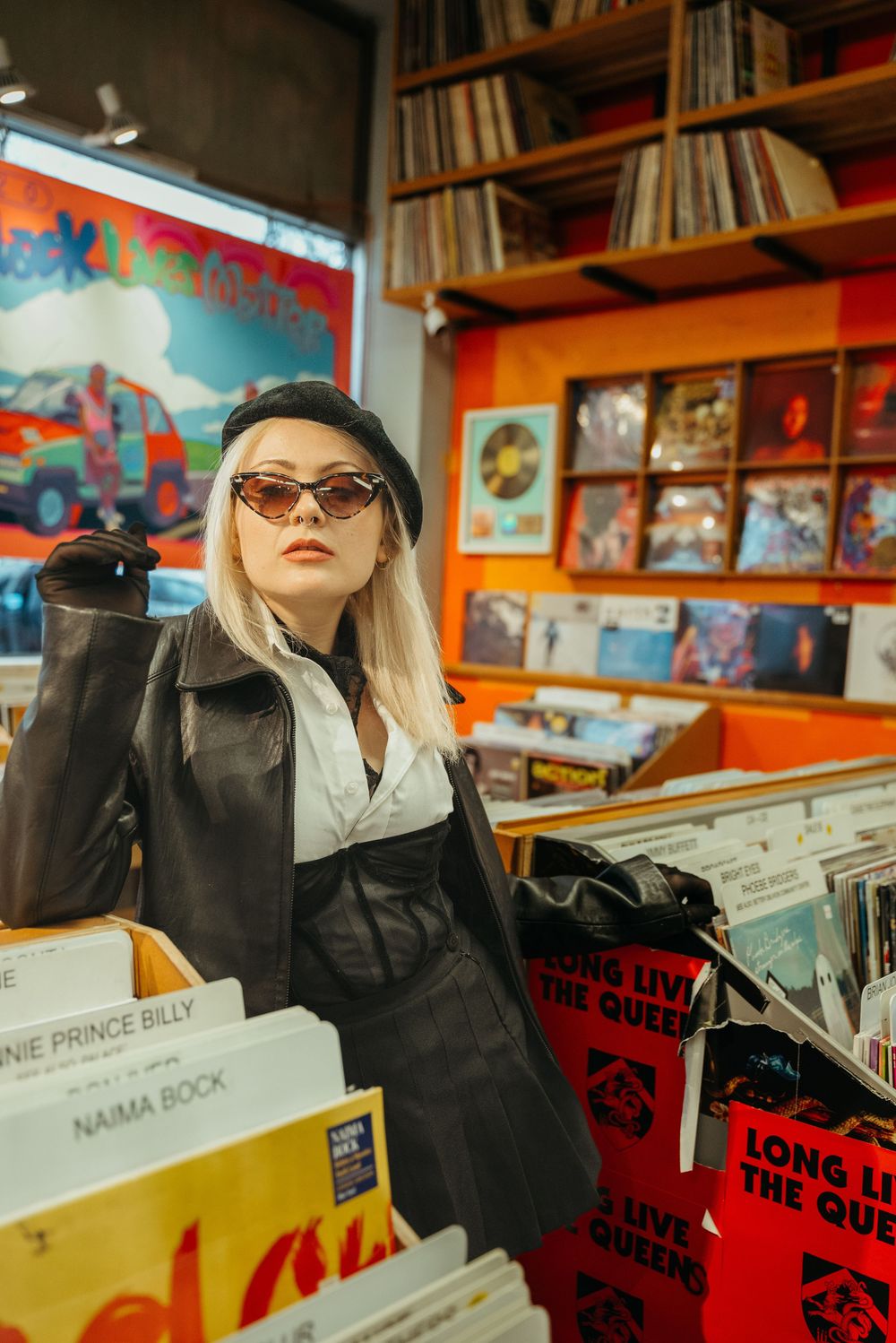 A stylish figure browses vinyl records in a record store wearing sunglasses and a black leather jacket.
