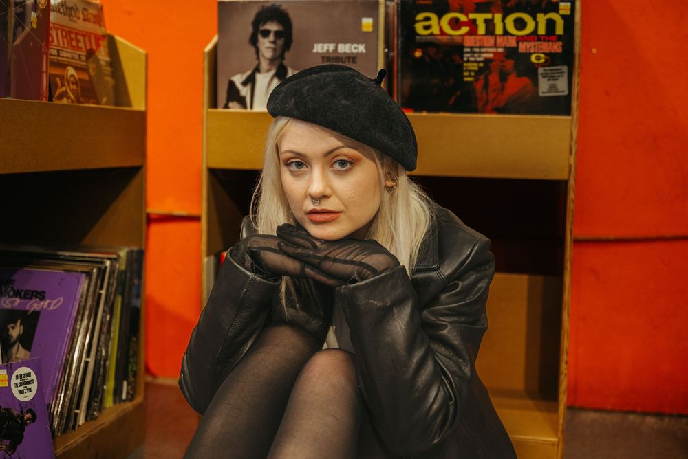 A person in a black leather jacket and beret sits crouched near vintage record albums against an orange wall.