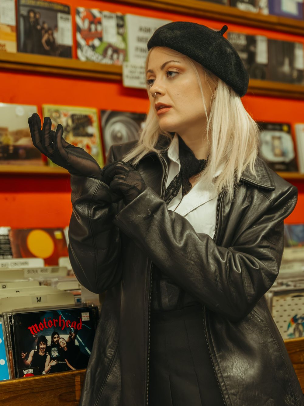 Model wearing black leather jacket and hat poses dramatically against orange backdrop with vinyl records.