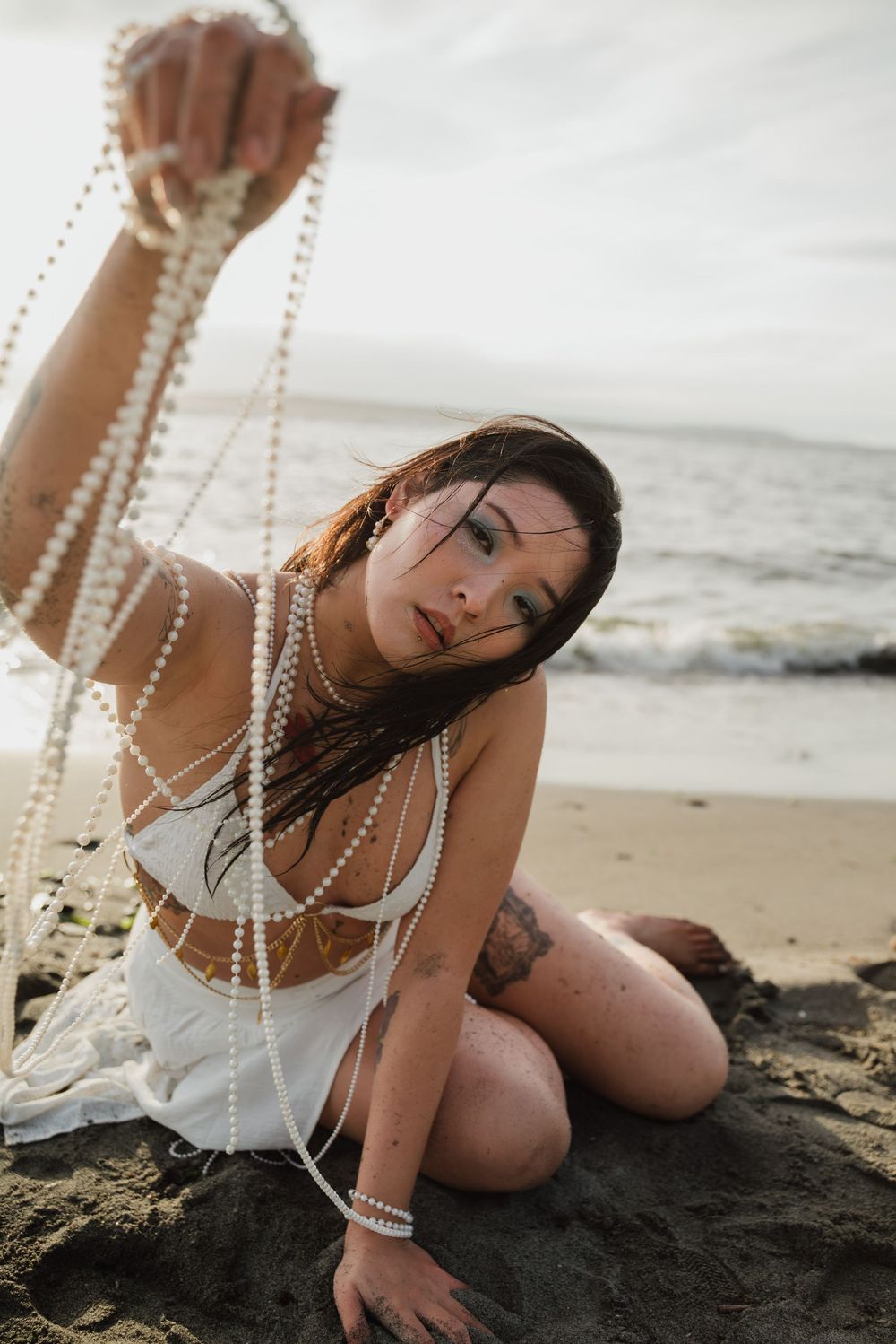 A person in a white dress poses with pearl necklaces on a beach at sunset.