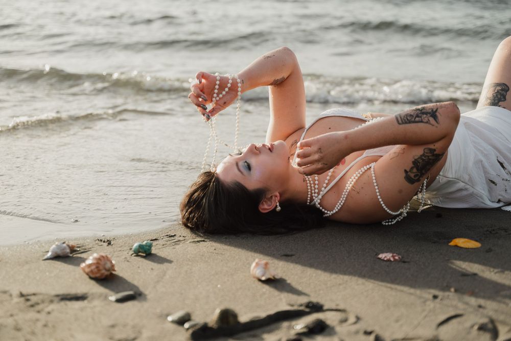 Person in white swimwear lying on sandy beach at sunset while seashells scatter nearby.