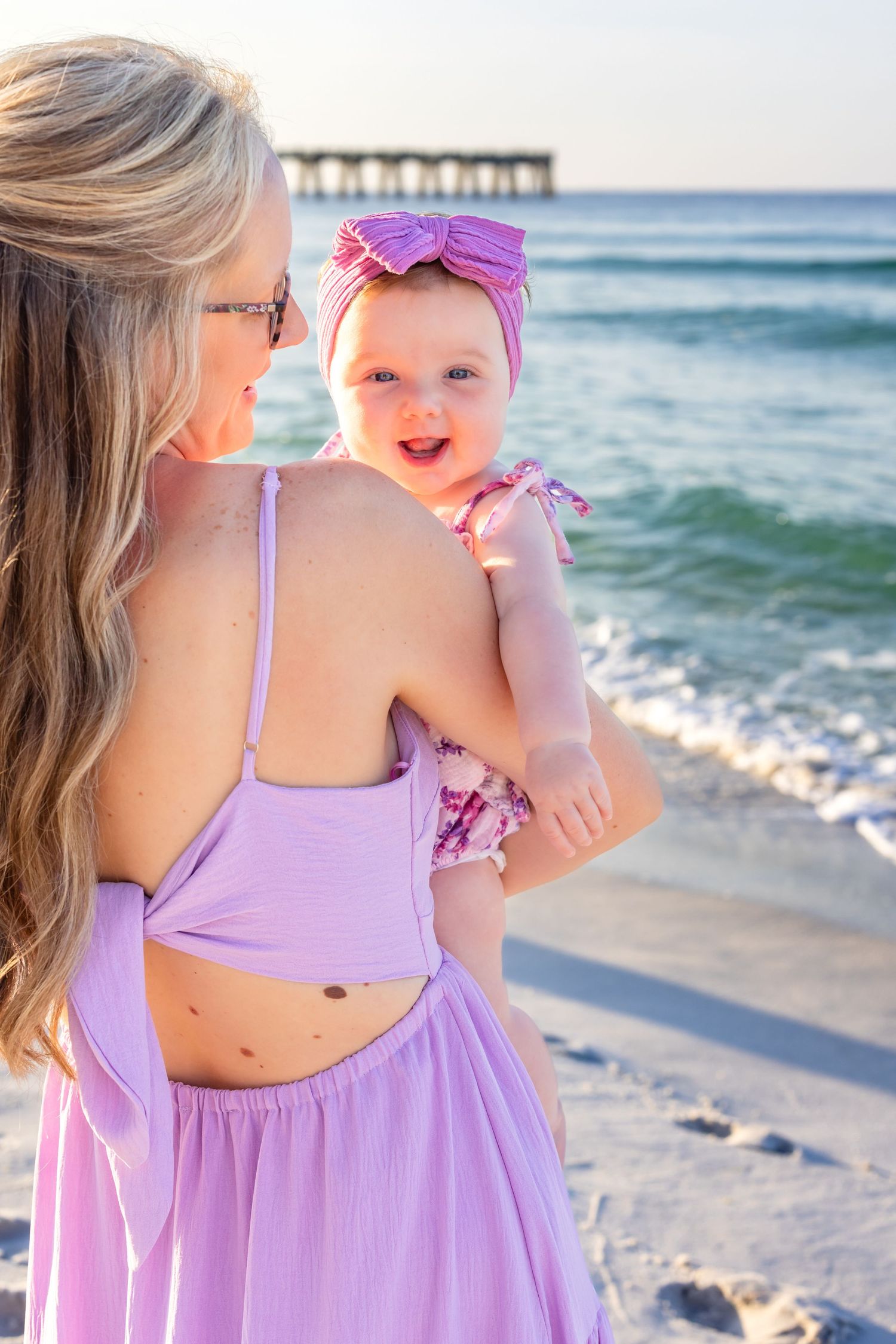 Mother and baby wearing matching purple outfits enjoy a sunny day at a beach pier with ocean waves in background.