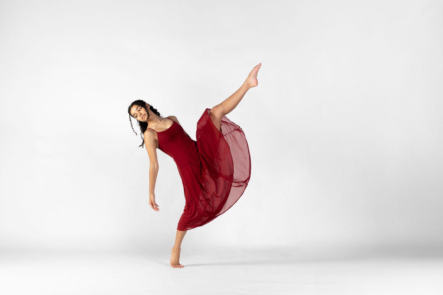 A dancer in a flowing red dress performs a graceful high kick against a white background.
