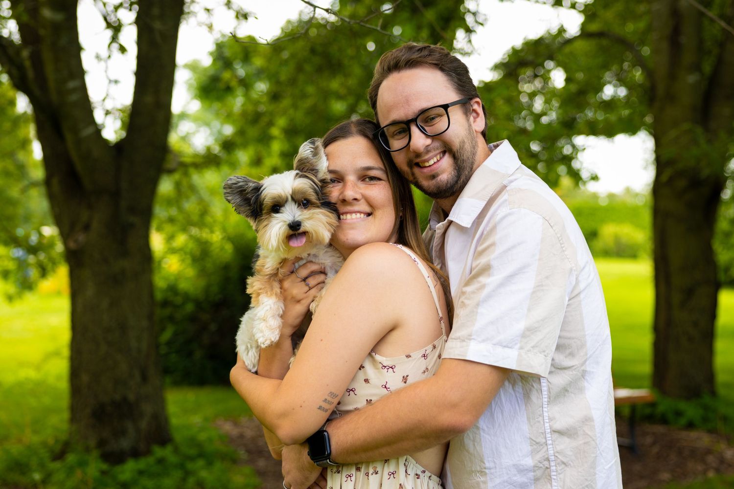 Couple in wedding attire embraces among trees while holding their small white dog.