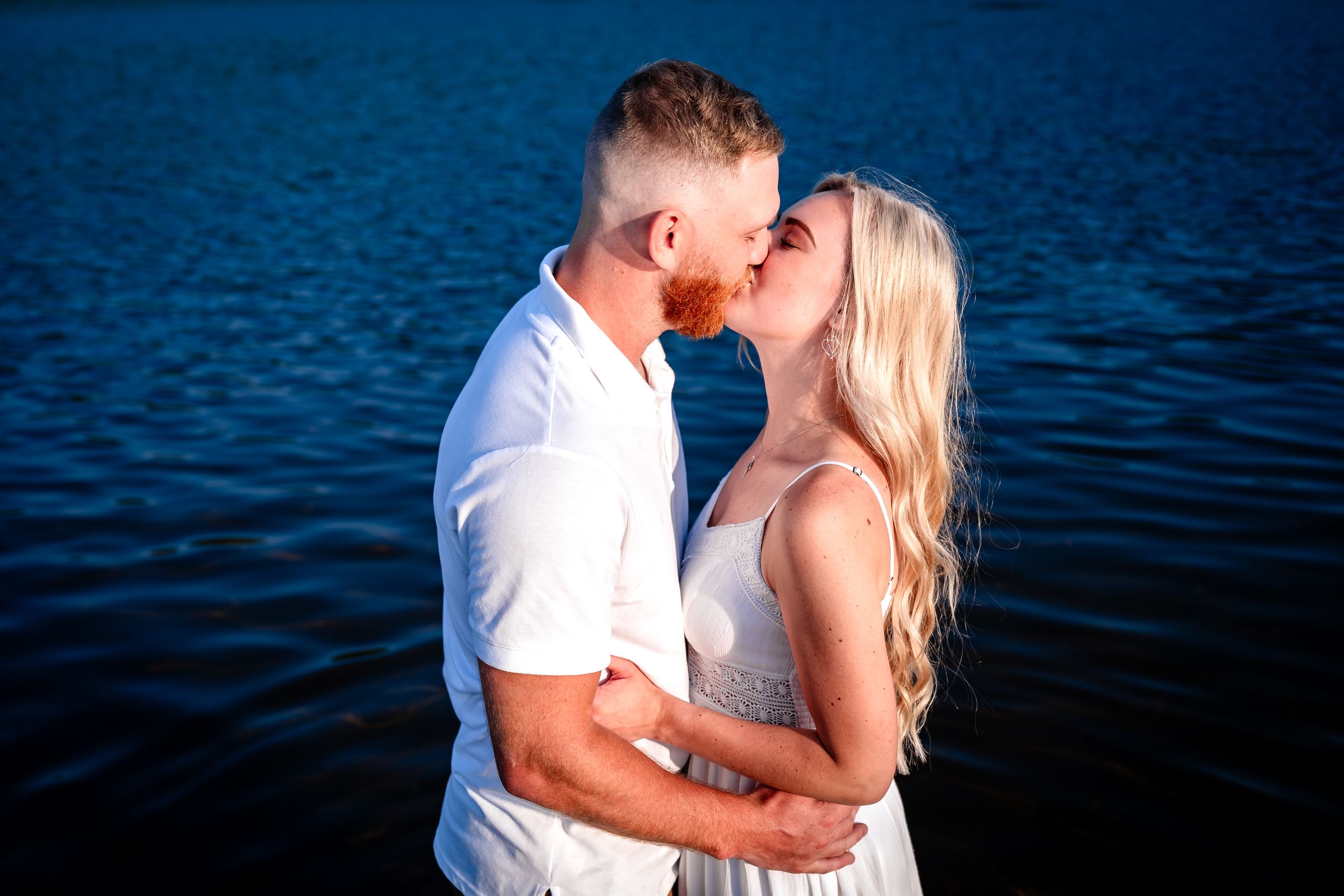 Couple embraces while standing in blue lake water during sunset photo session.