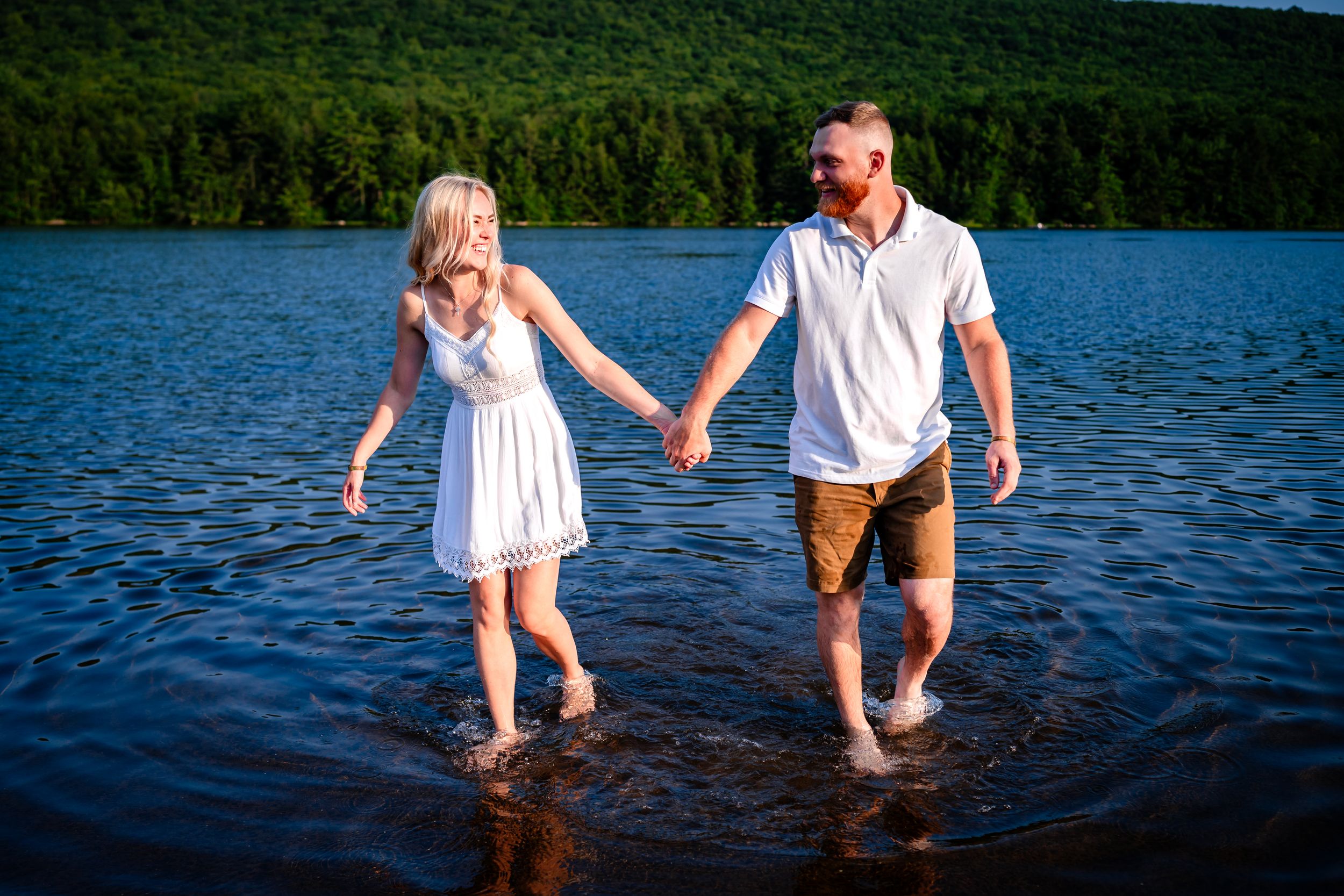Playful couple splashes in lake water wearing white outfits against forest background.