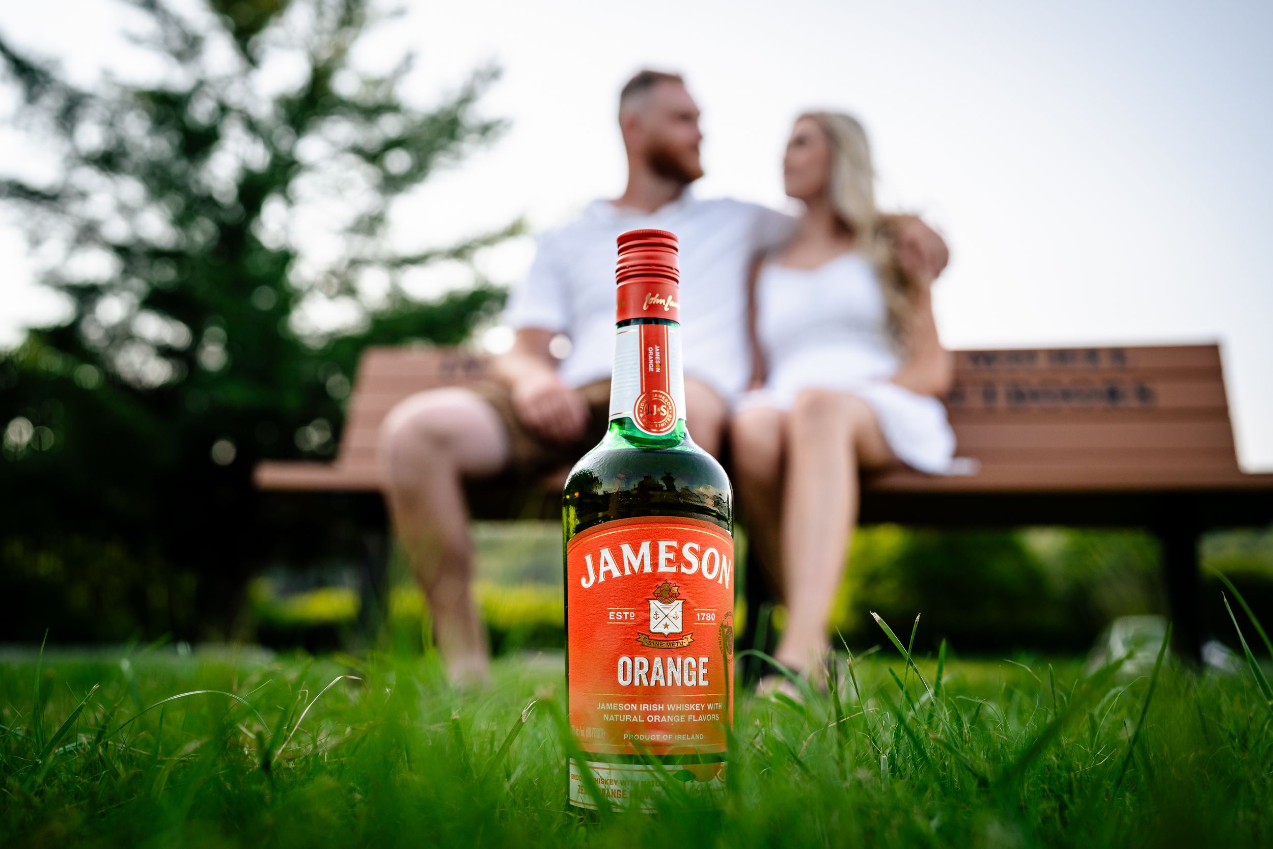 Jameson whiskey bottles displayed on grass with couple in soft focus background.