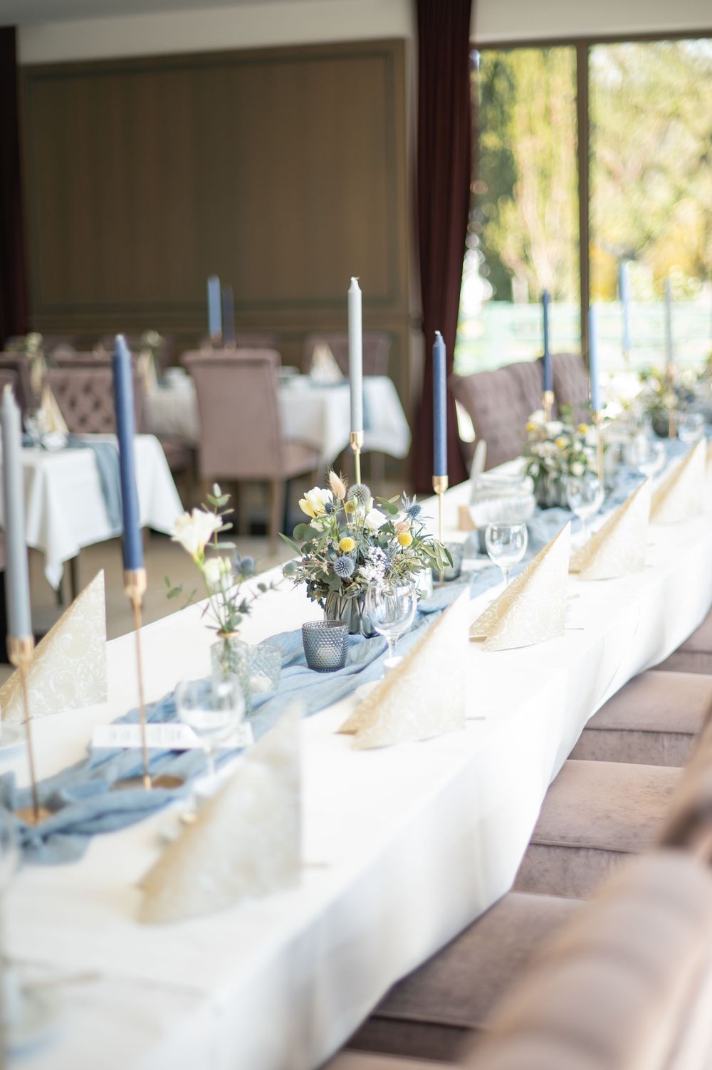 Elegant gedeckte Hochzeitstische mit blauen Kerzen und weißer Tischdecke in einem lichtdurchfluteten Raum.