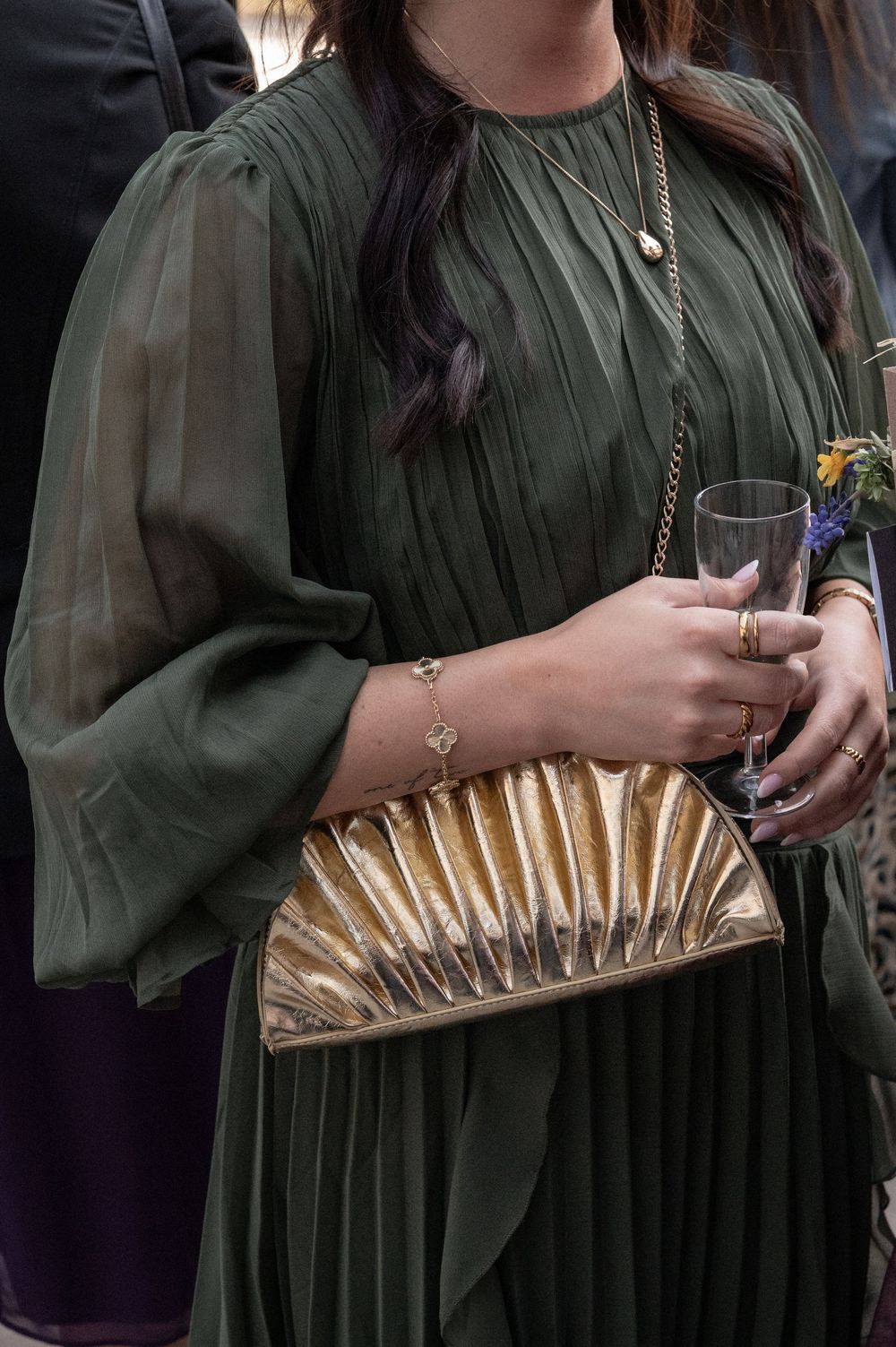 Eine goldfarbene, plissierte Clutch-Tasche in Metallic-Optik bildet einen eleganten Kontrast zu einem dunkelgrünen Kleid.