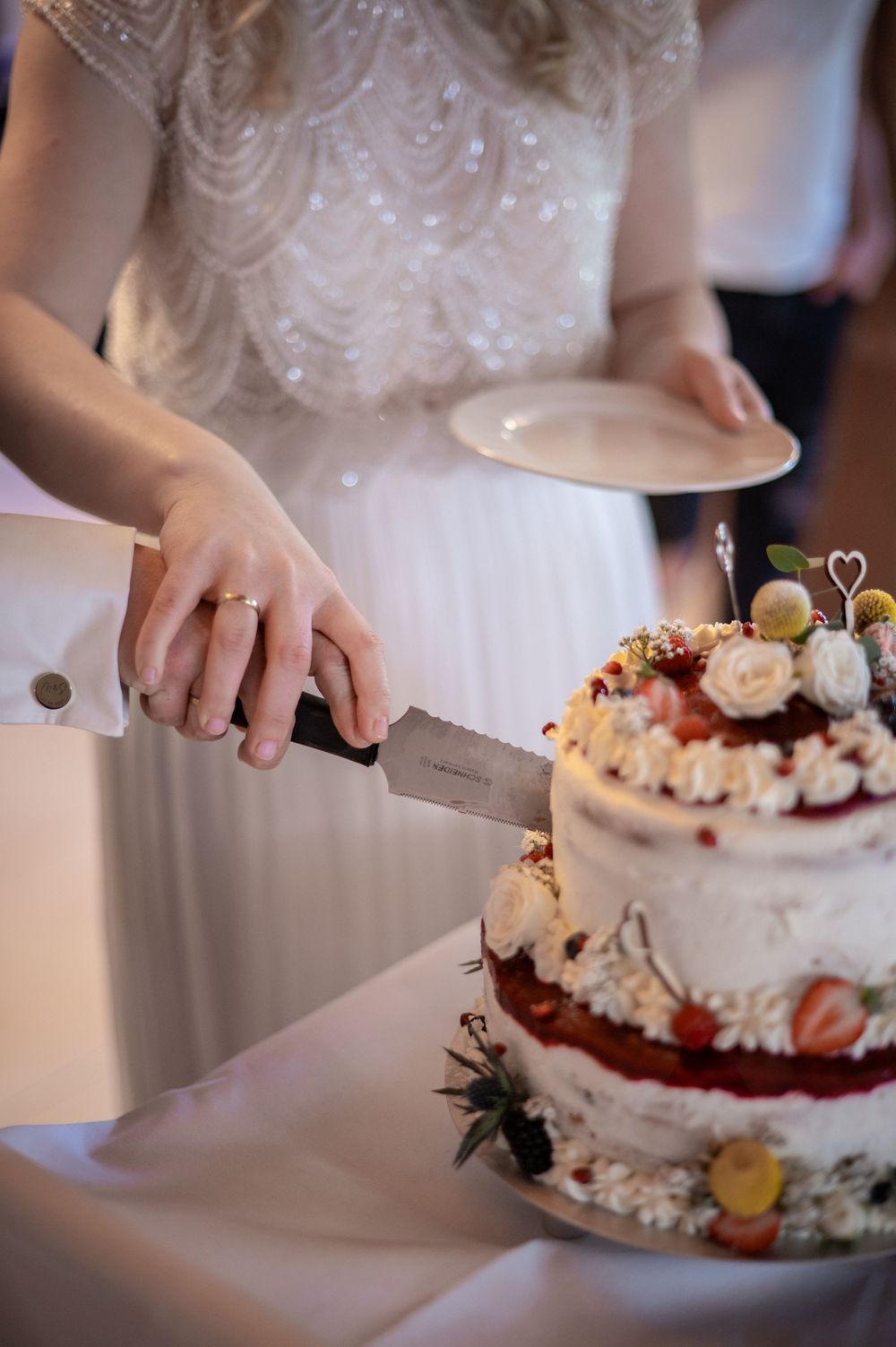 Die Braut schneidet in eine rustikale Hochzeitstorte, die mit frischen Blumen und Beeren verziert ist.