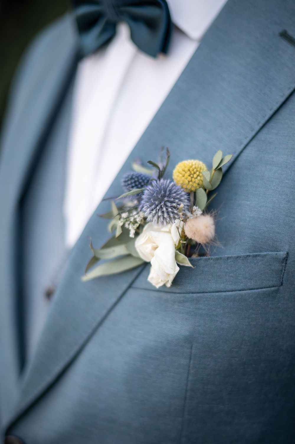 Nahaufnahme eines blaugrünen Anzugrevers mit einer eleganten Boutonniere aus weißen Rosen, gelben Craspedia und blauen Distelblüten.