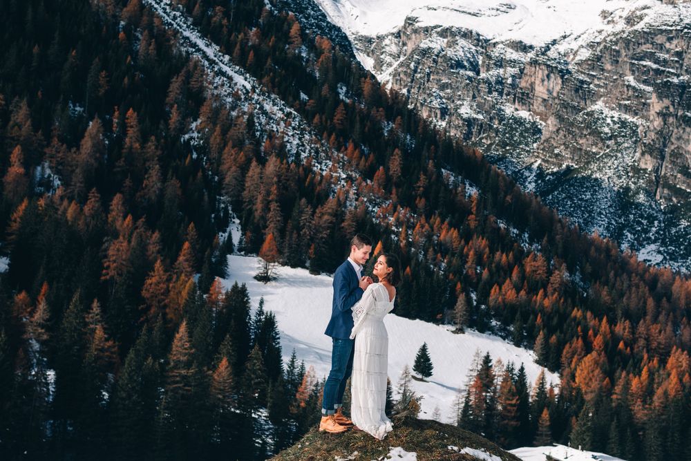 Two people stand on a cliff edge overlooking a snowy mountain valley with autumn larches and evergreens.