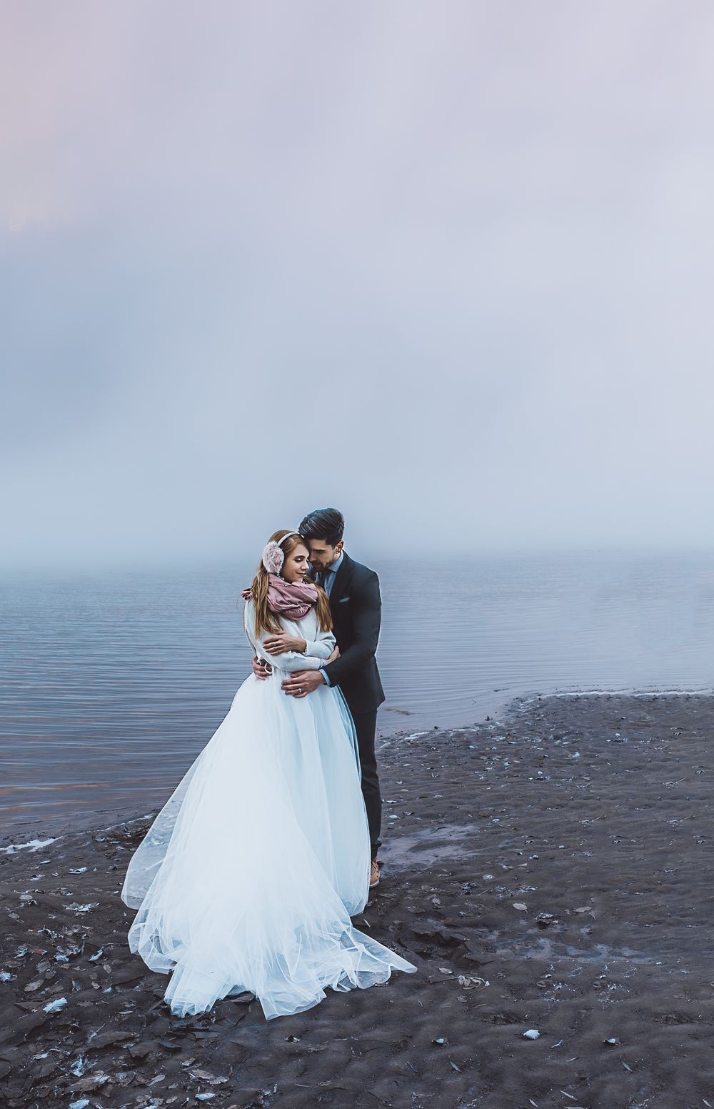 A romantic wedding couple embraces on a misty beach shoreline with calm waters and grey skies in the background.