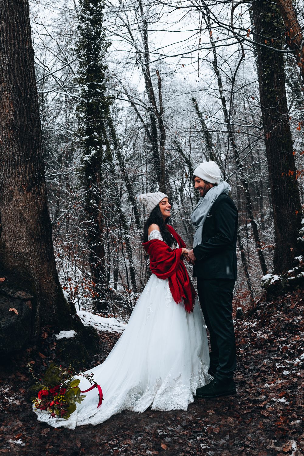 A winter wedding scene in a snowy forest with a bride in white dress and red wrap standing with partner among bare trees.