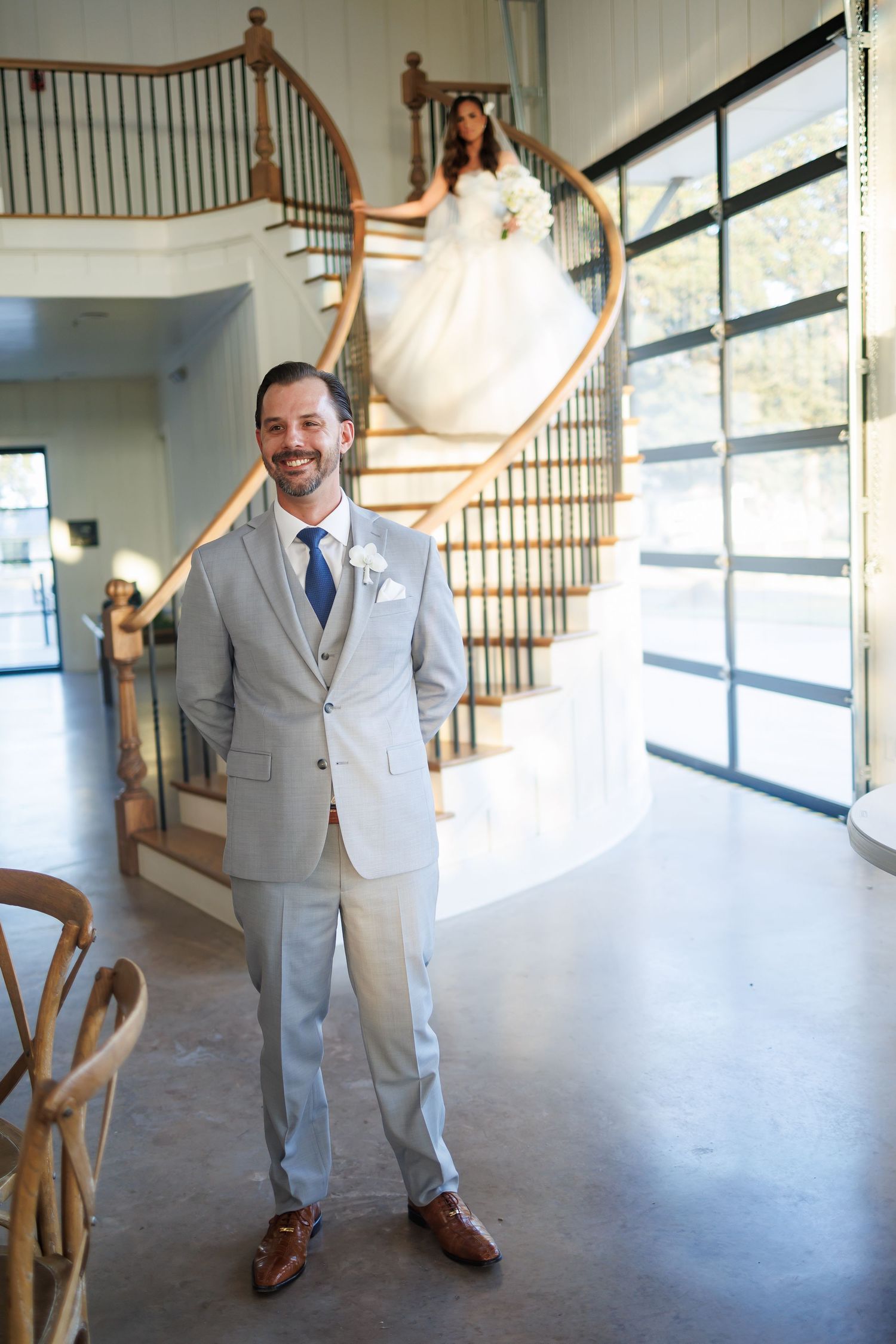 Wedding first look on elegant curved staircase at Bella Cavalli with large windows, bride in flowing white gown holds white floral bouquet.