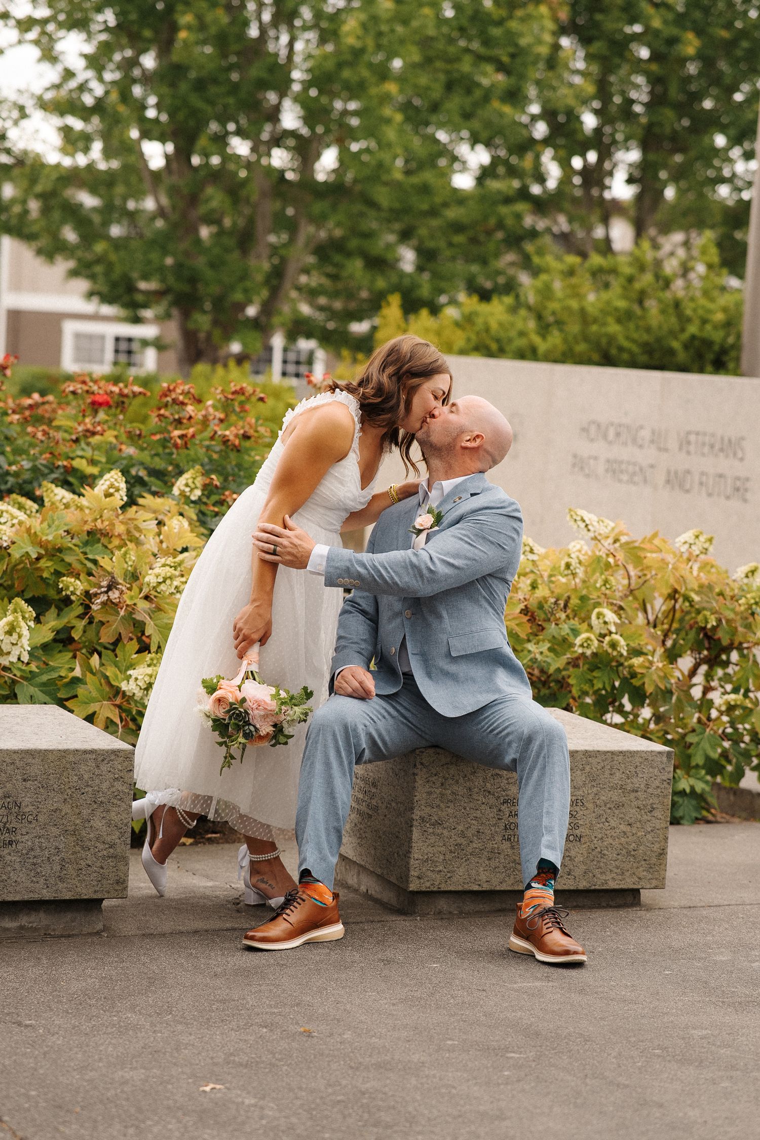 A romantic wedding couple sharing an intimate moment on a stone bench surrounded by flowers and greenery.
