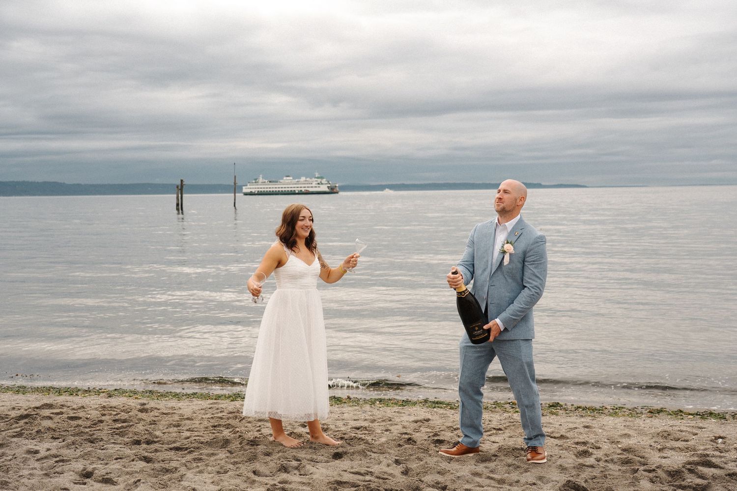 A wedding couple celebrates on a beach with a ferry boat visible in the distance on a cloudy day.