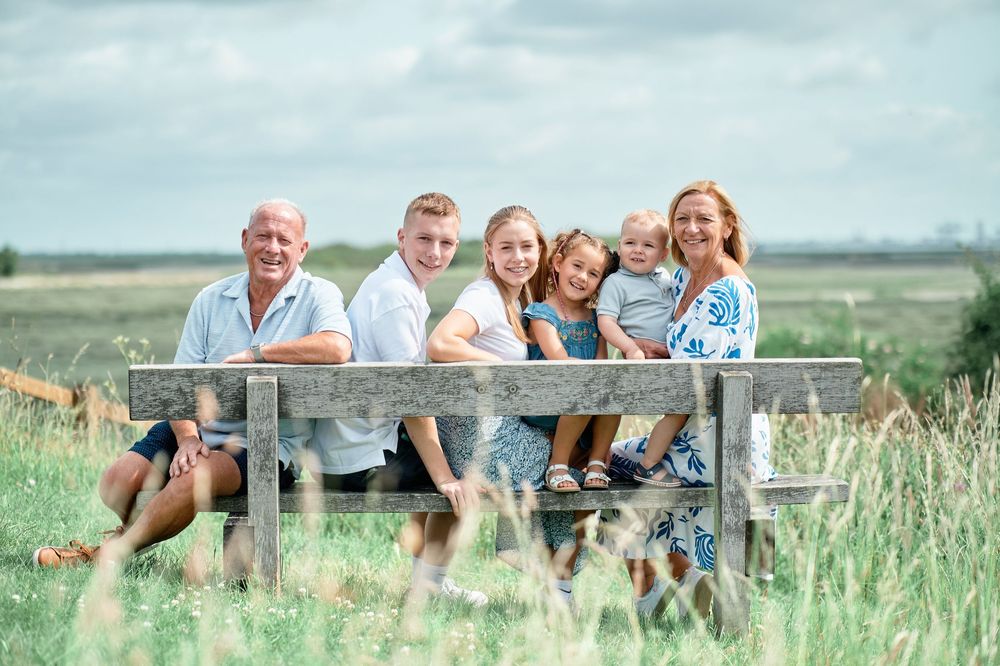 A group sits together on a wooden bench surrounded by tall grass in a sunny rural landscape.