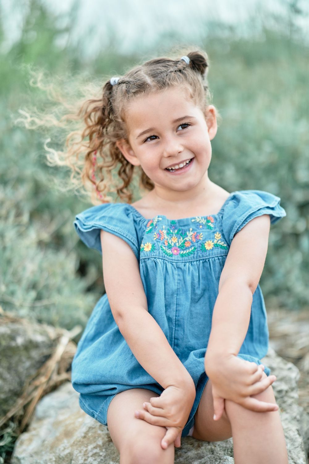 Child in blue embroidered dress sitting on rock outdoors with natural bokeh background.