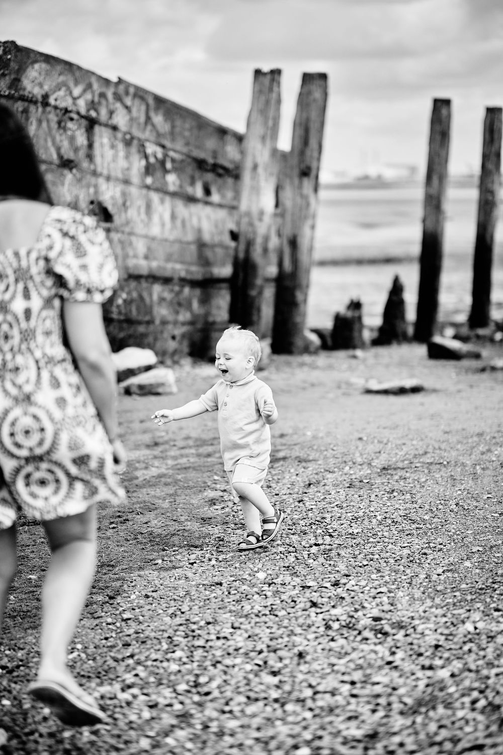 A black and white seaside scene shows a toddler walking on a pebbly beach near weathered wooden posts under cloudy skies.