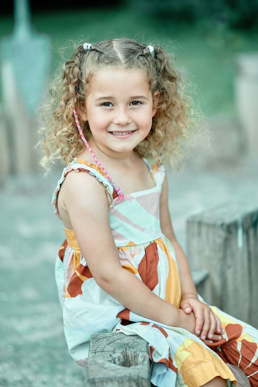 Child with curly hair wearing colorful sundress sits outdoors in natural setting with soft teal background.