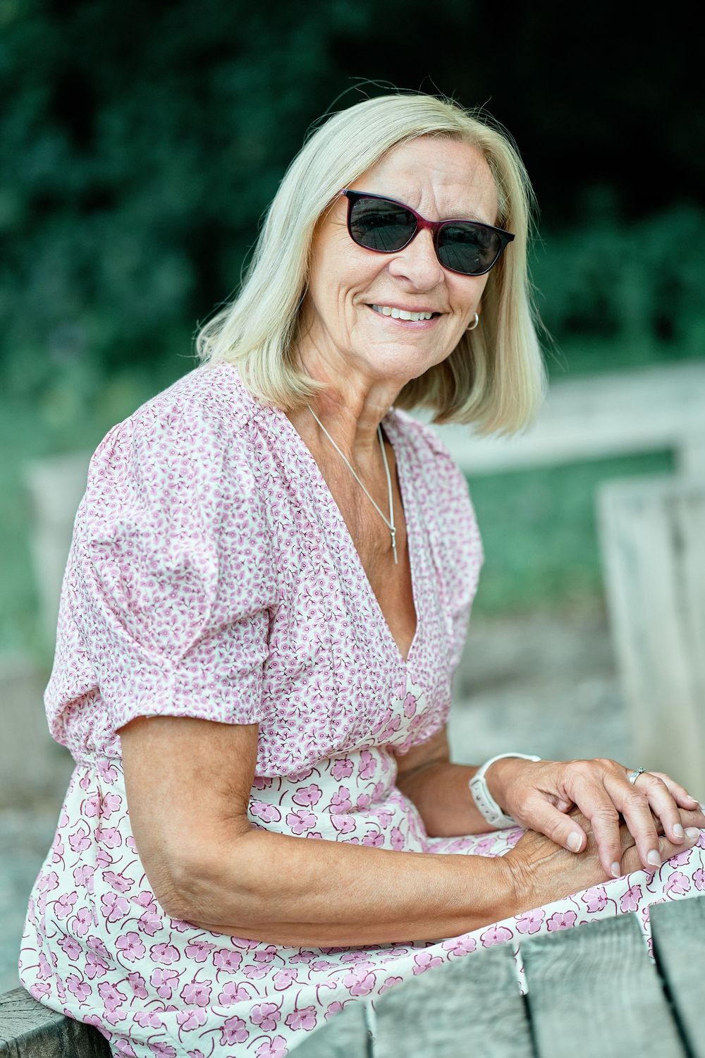 Woman in floral pink dress and sunglasses relaxes on wooden surface outdoors on sunny summer day.