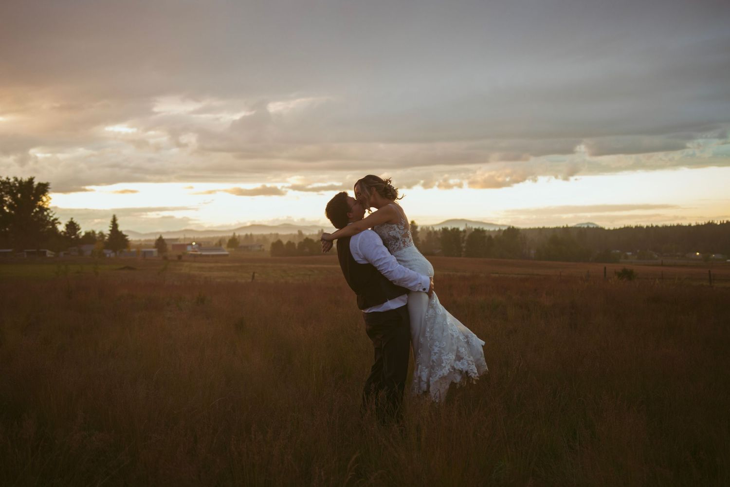A couple stands in a sunlit meadow wearing wedding attire surrounded by evergreen trees during golden hour.