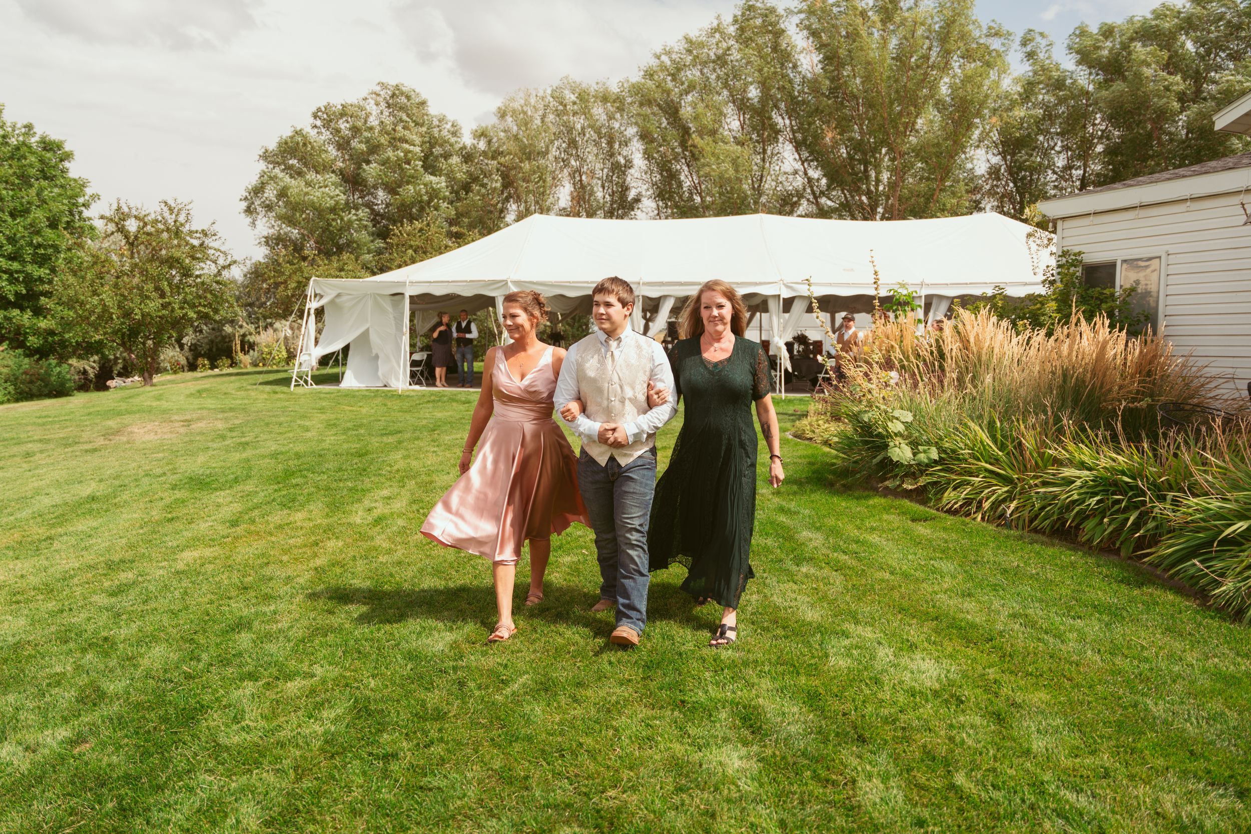 A series of people walking across a sunny green lawn with trees in the background.