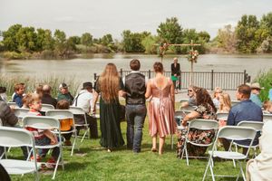 An outdoor waterfront wedding ceremony with guests seated in white folding chairs by a lake.