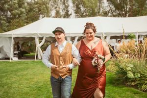 A couple walks joyfully past a white tent at an outdoor wedding reception.