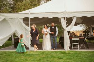 Wedding party members gather under a white tent on a grassy lawn during a celebration.