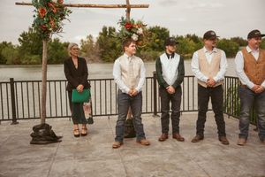 Wedding party members stand in a line on a deck overlooking water under a floral arch.
