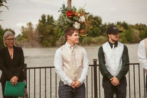 Wedding party members stand at attention under a rustic wooden arch decorated with flowers.