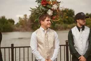A groomsman stands under a rustic wooden arch decorated with orange and white flowers by a waterfront.