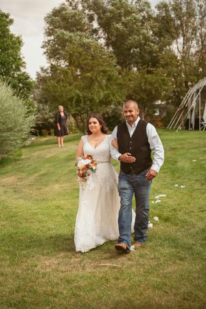 A wedding couple walks across a grassy lawn with trees and a white tent in the background.