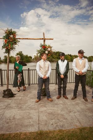 A wedding party stands under a wooden cross arch decorated with orange and green floral arrangements by a waterfront.