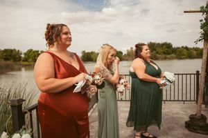 Wedding party in colorful formal dresses stands at a lakeside venue during golden hour.