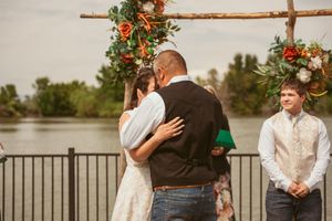 A romantic wedding ceremony moment under a rustic wooden arch decorated with colorful flowers by a lake.