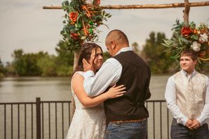 A couple embraces during their wedding ceremony under a wooden arch decorated with orange flowers.
