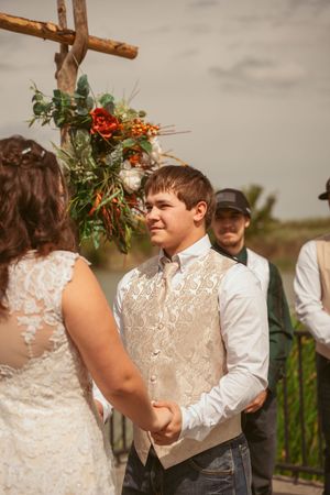 A couple holds hands during their rustic outdoor wedding ceremony beneath a cross decorated with flowers.
