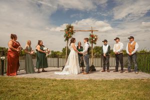 An outdoor lakeside wedding ceremony with attendants in earth tones gathered under a wooden cross arch.