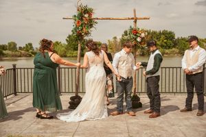 Wedding ceremony takes place under a wooden arch decorated with orange and green florals by a waterfront.