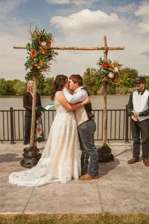 A couple shares their first kiss under a wooden arch decorated with orange and white flowers.