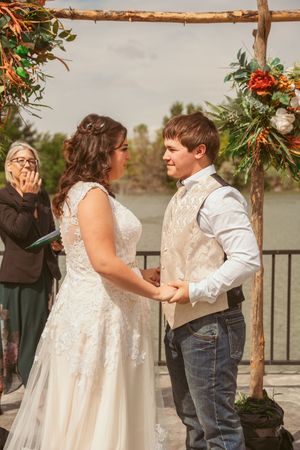 A couple exchanges vows under a rustic wooden arch decorated with flowers during an outdoor ceremony.