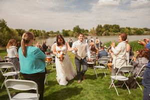 Wedding guests stand and applaud as newlyweds walk down the aisle on a grassy lawn by a lake.