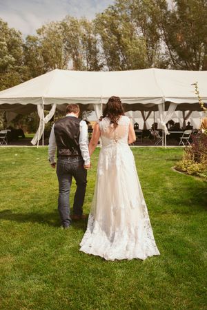A couple walks across a lawn towards an outdoor wedding reception tent on a sunny summer day.